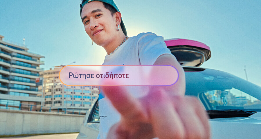 Νεαρός άνδρας χαμογελάει και δείχνει προς την κάμερα, προσκαλώντας σε διάδραση. Μπροστά του, ένα κουμπί αναγράφει «Ρώτησε οτιδήποτε».